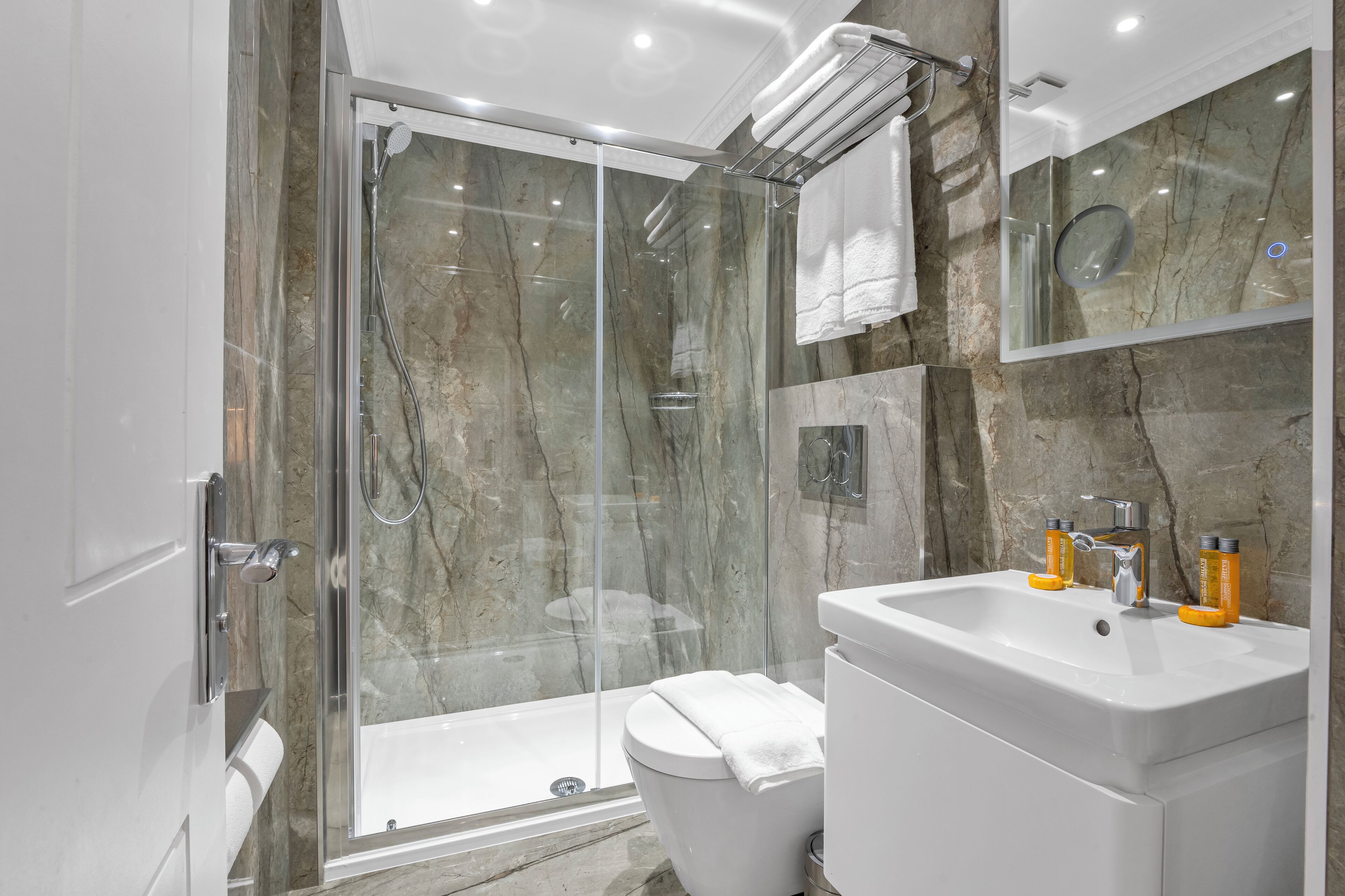 A modern bathroom with a glass-enclosed shower, a white sink with two orange soap dispensers, and a marble-tiled wall.
