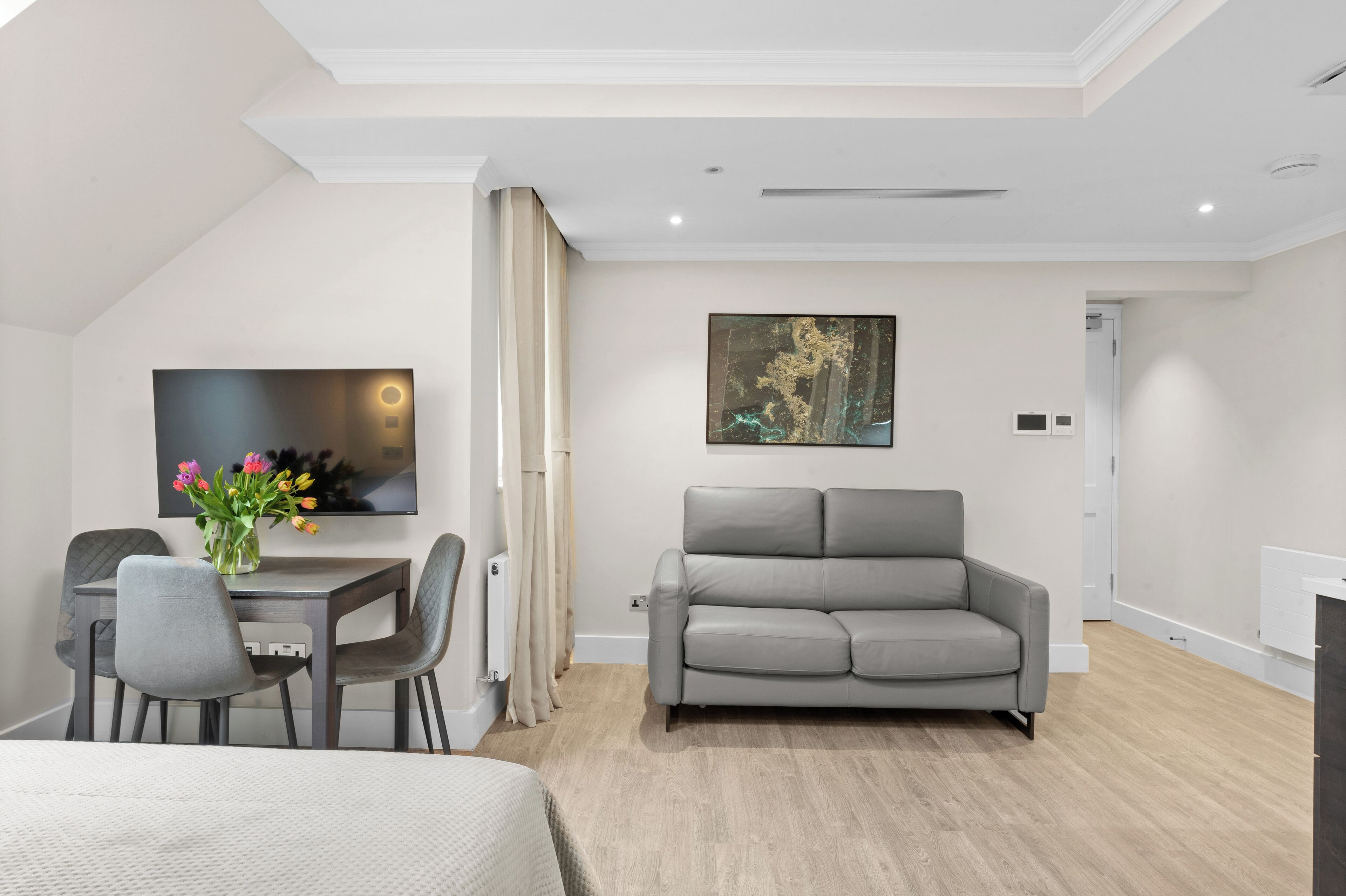 Una habitación de hotel moderna con una cama, un sofá gris, una mesa de comedor con sillas, un televisor de pantalla plana y un cuadro en la pared.