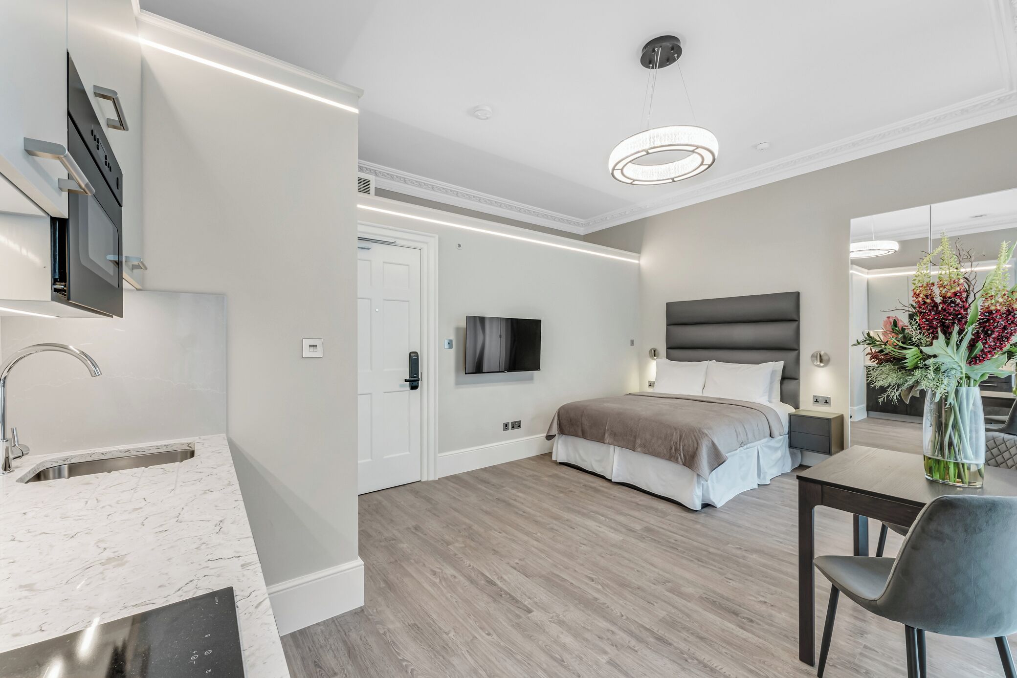 A modern hotel room with a kitchen area, a large bed, a bedside table, and a mirror.