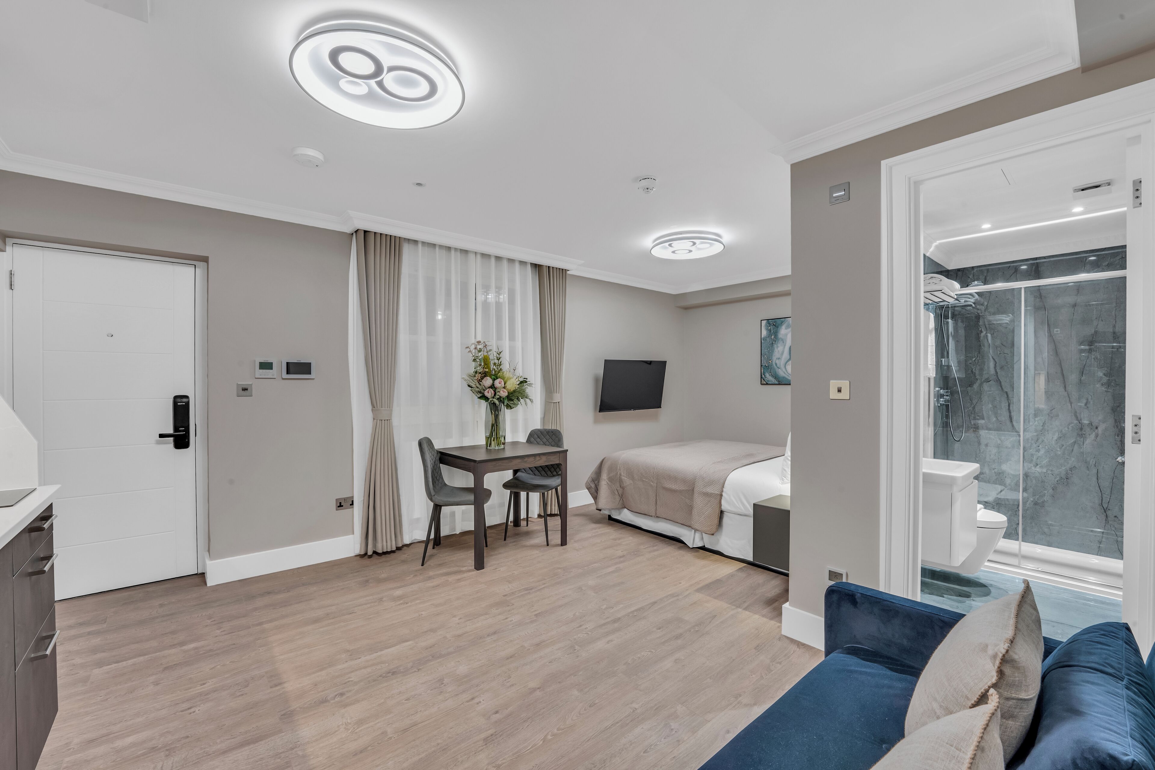 A modern hotel room with a dining area, a bed, a TV, and a shower.