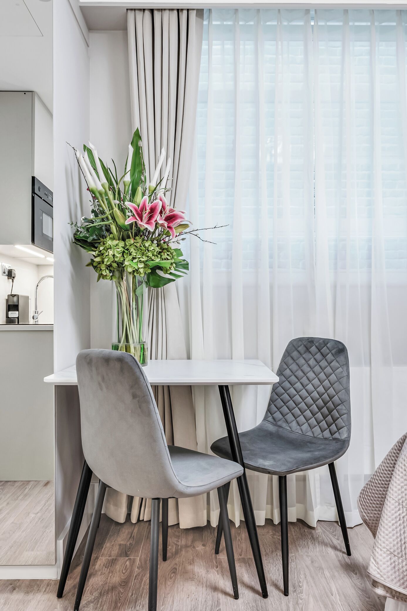 A modern dining area with a white table, two chairs, and a floral centerpiece.
