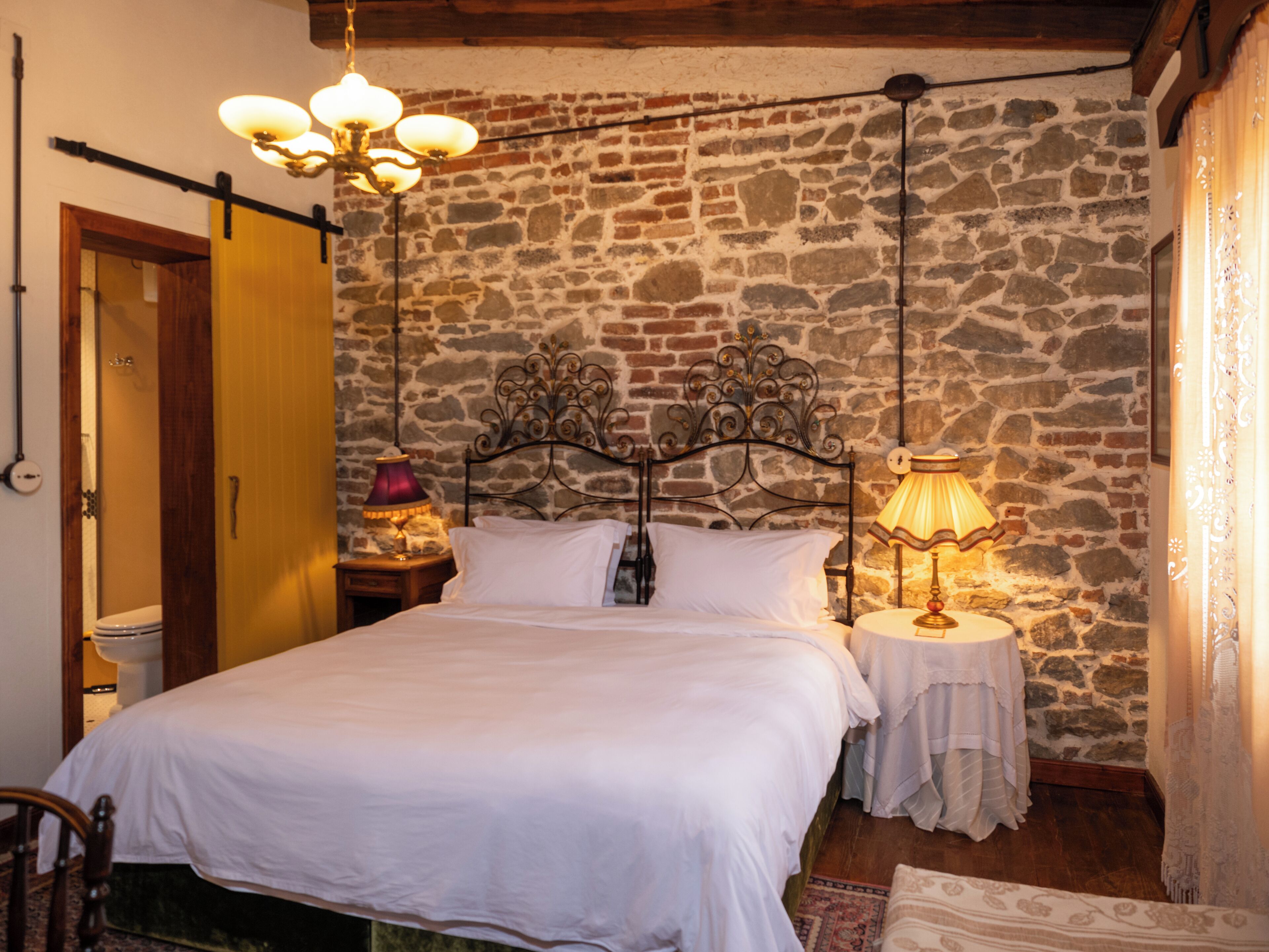 A bedroom with a stone wall, a bed with white linens, two bedside lamps, and a wooden door.