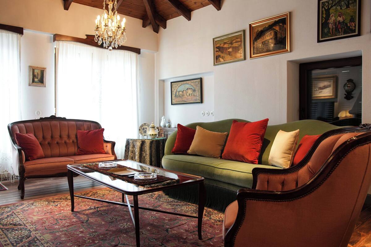 Ein Wohnzimmer mit Sofa, Sessel und Couchtisch. An den Wänden befinden sich Gemälde und eine Deckenleuchte.
