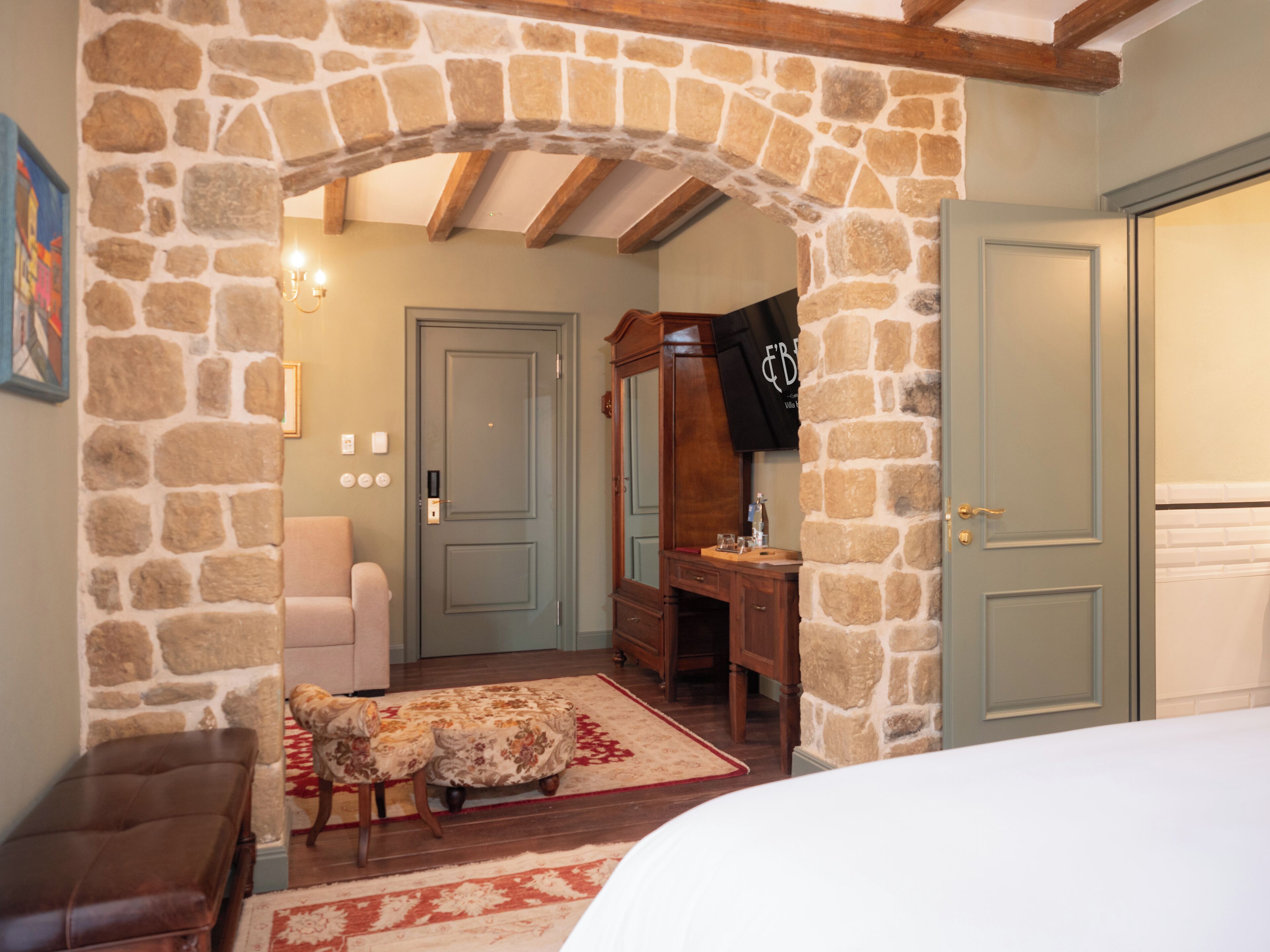 Une pièce dotée d’une arche en pierre, d’un banc en bois, d’une petite table recouverte d’une nappe à motifs floraux et d’une porte à laquelle est accroché un sac noir.