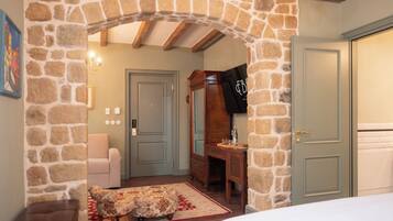 Une pièce dotée d’une arche en pierre, d’un banc en bois, d’une petite table recouverte d’une nappe à motifs floraux et d’une porte à laquelle est accroché un sac noir.