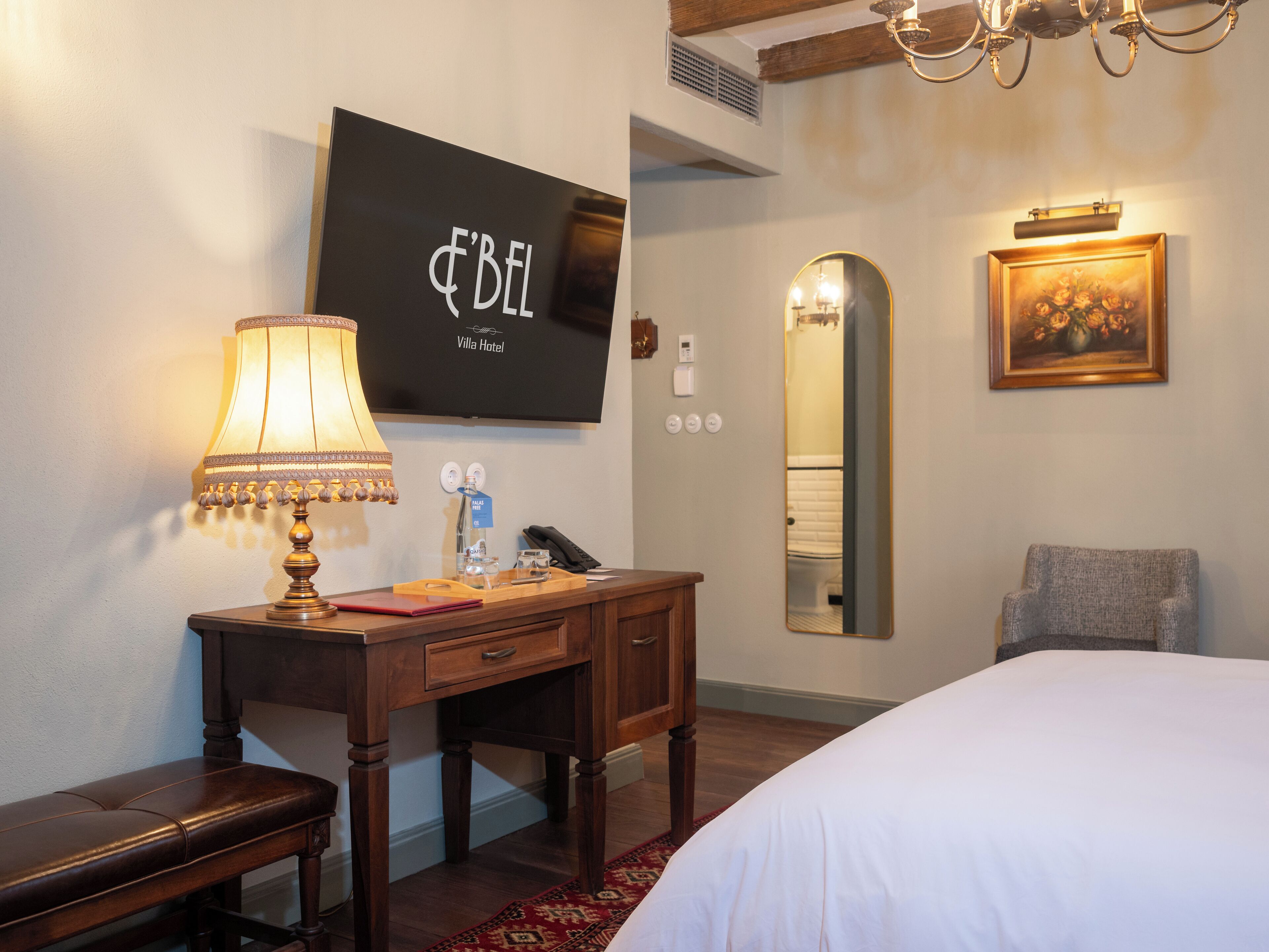 Et hotelværelse med en seng, et skrivebord med lampe, et spejl, et maleri og et fjernsyn der viser "F'BEL Villa Hotel".