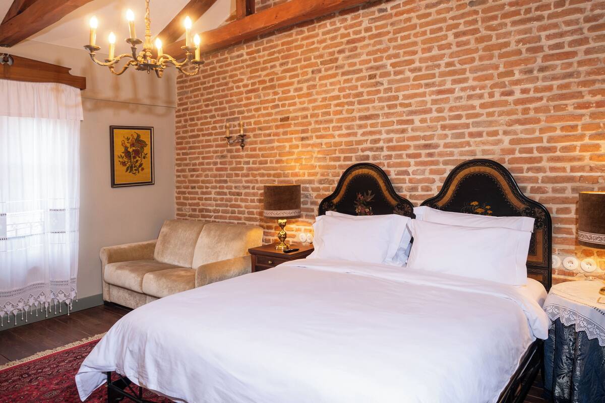 Ein Schlafzimmer mit Backsteinwand, einer Kandelaber, einem Bett mit weißen Leinen, einem Holz-Kopfteil, einem Nachttisch, einer Lampe und einem Fenster mit Vorhängen.