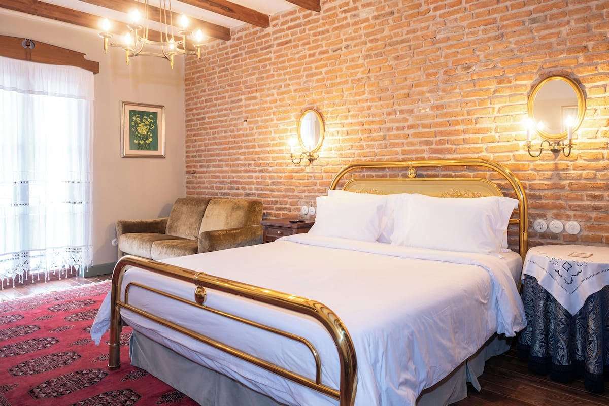 Ein Zimmer mit Backsteinwand, einem Bett mit goldener Rahmung, einer Holzbalkendecke und einem roten Teppich.