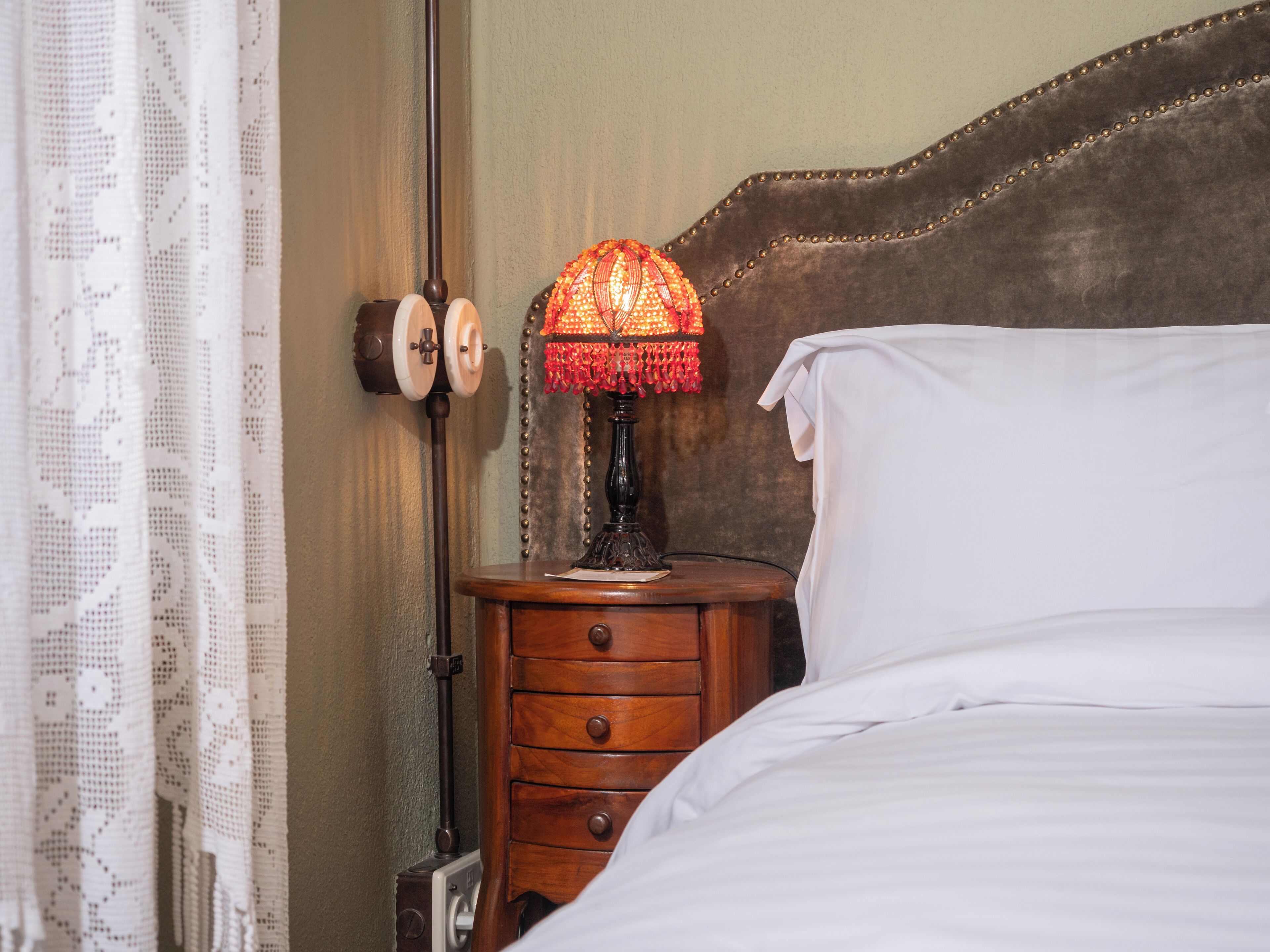 A neatly made bed with white linens, a wooden nightstand with a lamp, and a lace curtain.