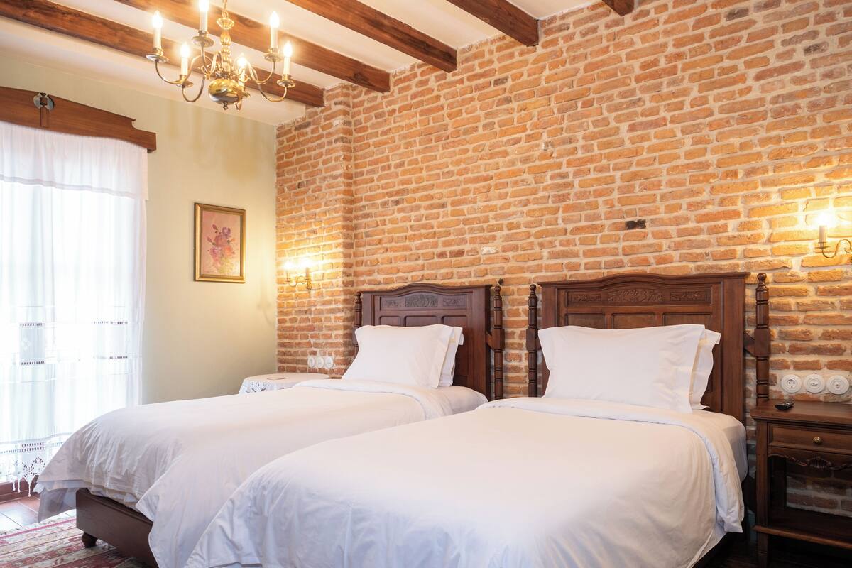 Ein Hotelzimmer mit zwei Betten, einer Ziegelwand, Holzbalken, einer Chandelüster und einem Nachttisch mit Lampe.