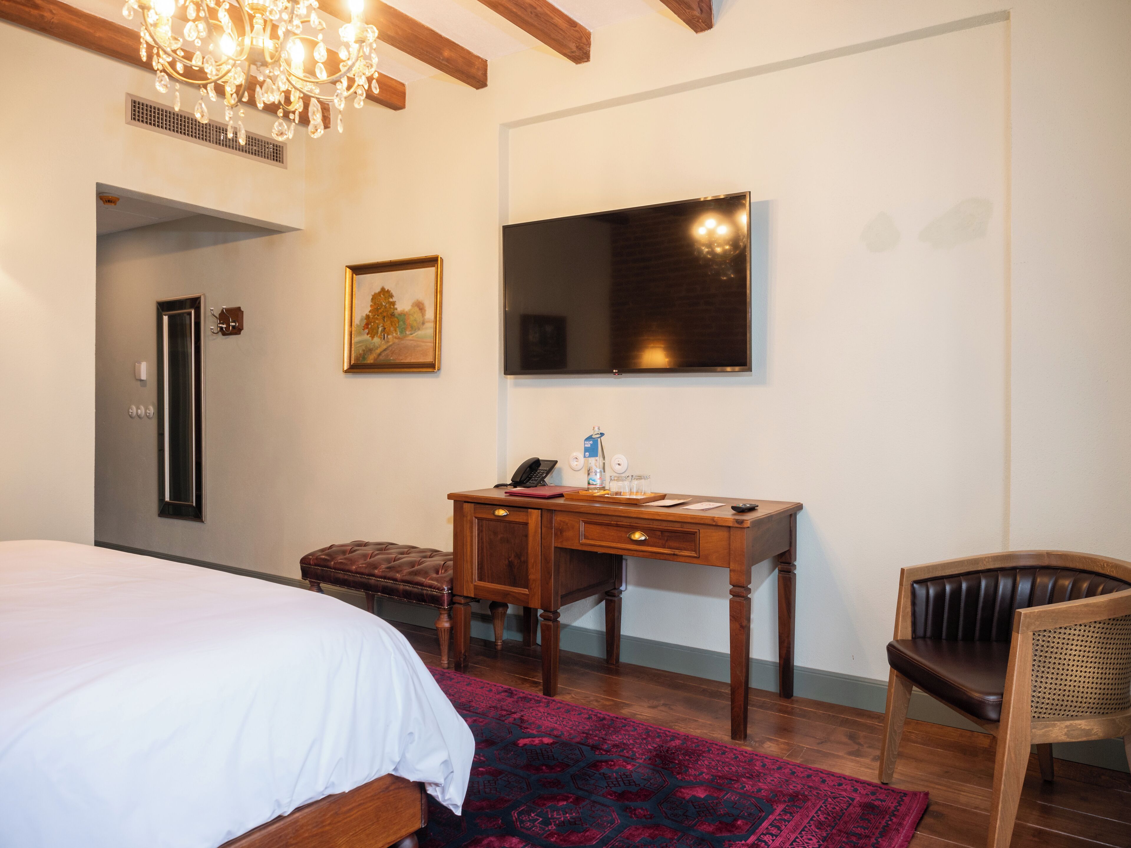 A hotel room with a bed, a desk with a telephone, a chair, a mirror, and a chandelier.