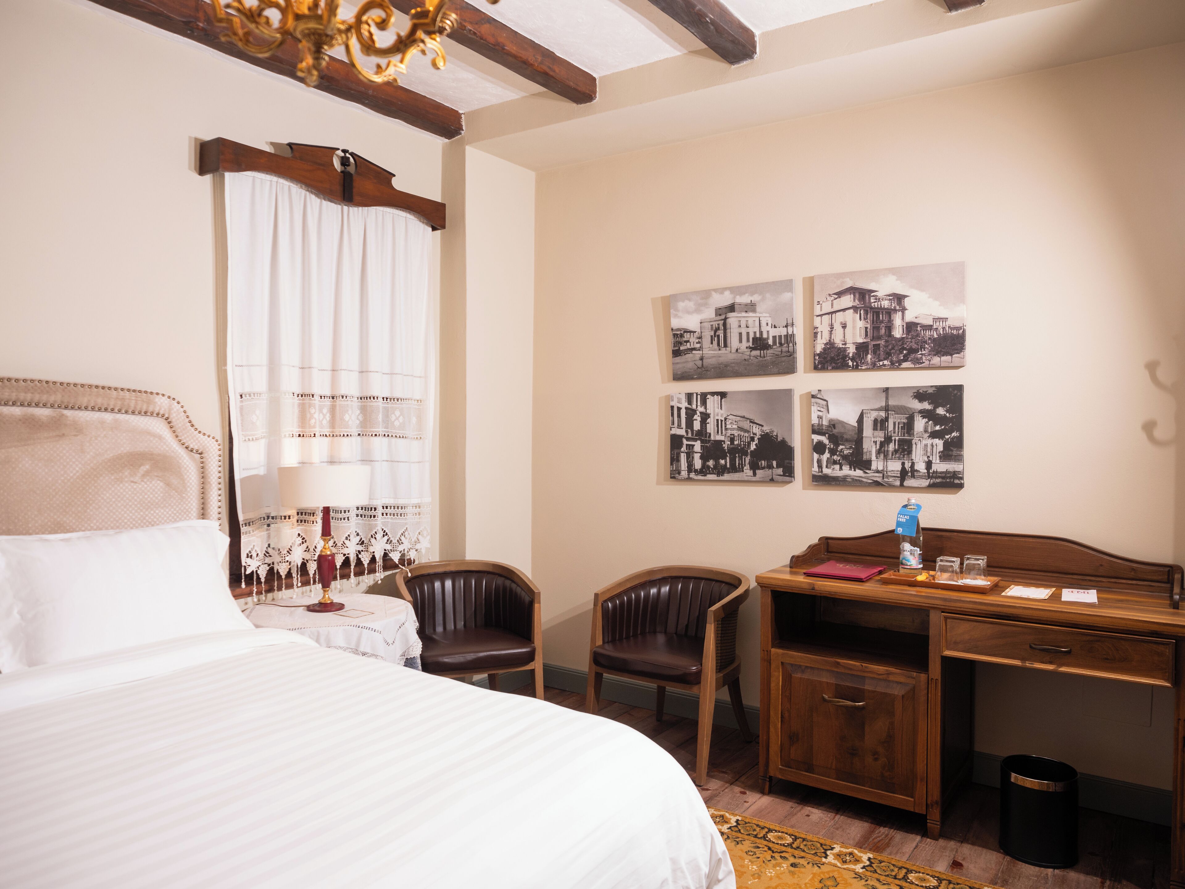 A hotel room with a bed, a desk, two chairs, a small table with a lamp, and a wall with four black-and-white photographs.