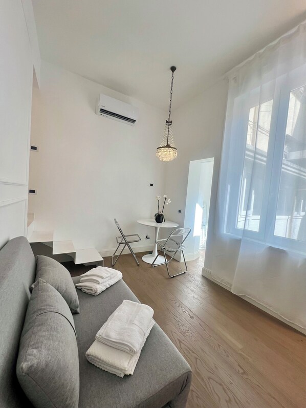 Ein modernes Wohnzimmer mit einem grauen Sofa, einem kleinen runden Tisch und zwei Stühlen. Eine Deckenleuchte hängt von der Decke, und ein Fenster mit transparenten Vorhängen ist zu sehen.