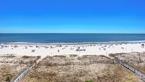 A beach with a wooden walkway leading to the water, sandy beach, and a flagpole with a flag. - Lighthouse 506 by Liquid Life (Gulf Shores)