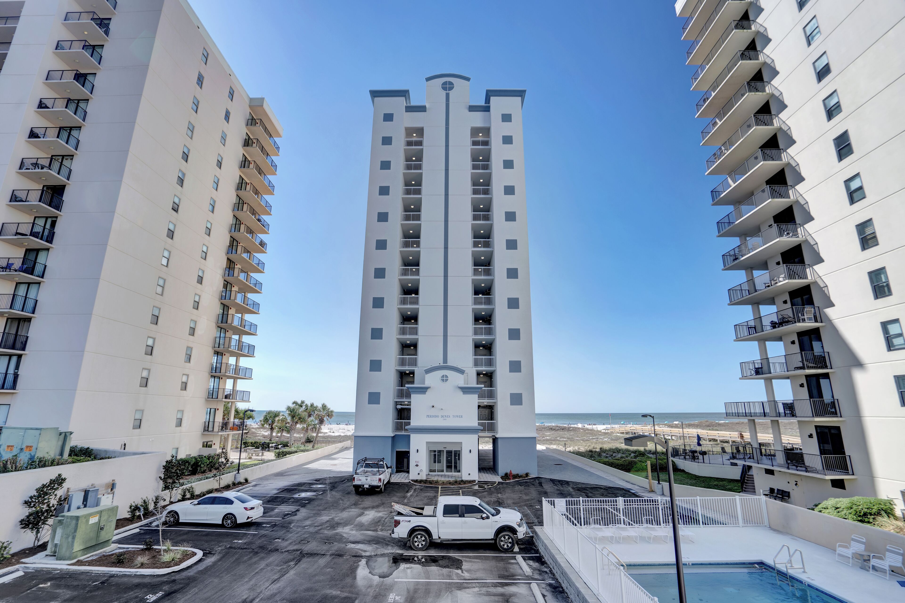 A high-rise hotel with a central entrance, parking lot, and a pool area.