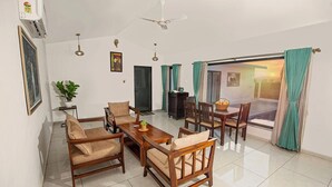 A modern living room with a sofa set, dining table, and chairs. There is a ceiling fan, a plant, and a window with curtains. - The Stay Ranakpur (Desuri)