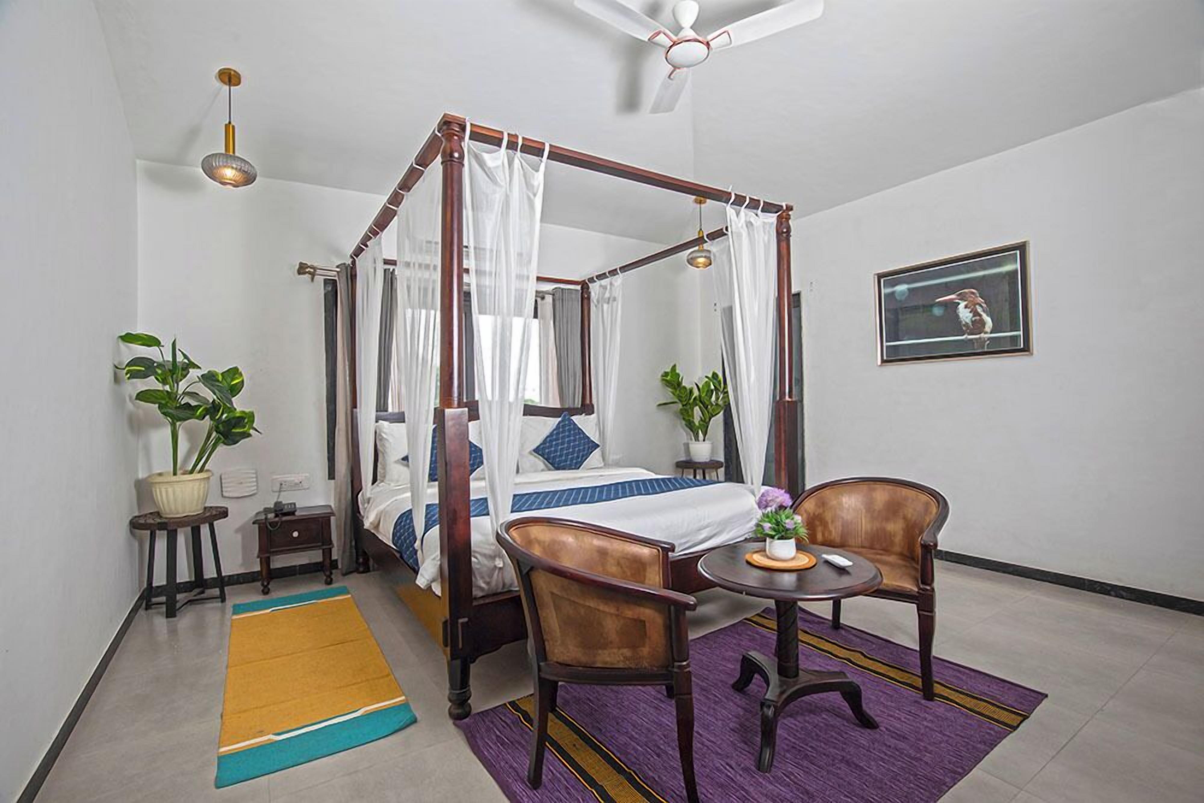 A four-poster bed with a canopy, a small round table with a chair, a potted plant, and a framed picture on the wall.