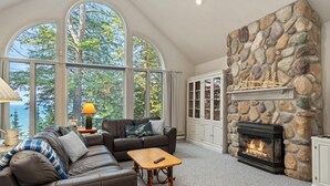 A cozy living room with a stone fireplace, large windows, and a comfortable seating area. - Lakeside Home with Panoramic Views of Lake Michigan! (Harbor Springs)