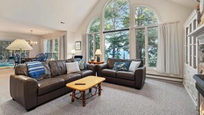 A spacious living room with a large window, a fireplace, and a pool visible through the window. - Lakeside Home with Panoramic Views of Lake Michigan! (Harbor Springs)
