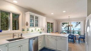 A kitchen with white cabinets, a dishwasher, and a dining area with blue chairs and a view of the ocean. - Lakeside Home with Panoramic Views of Lake Michigan! (Harbor Springs)