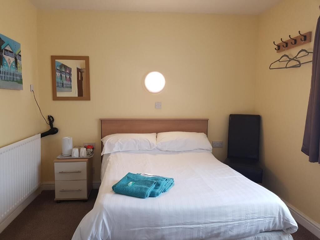 A hotel room with a bed, a bedside table with a lamp, a wall-mounted picture, and a coat rack.