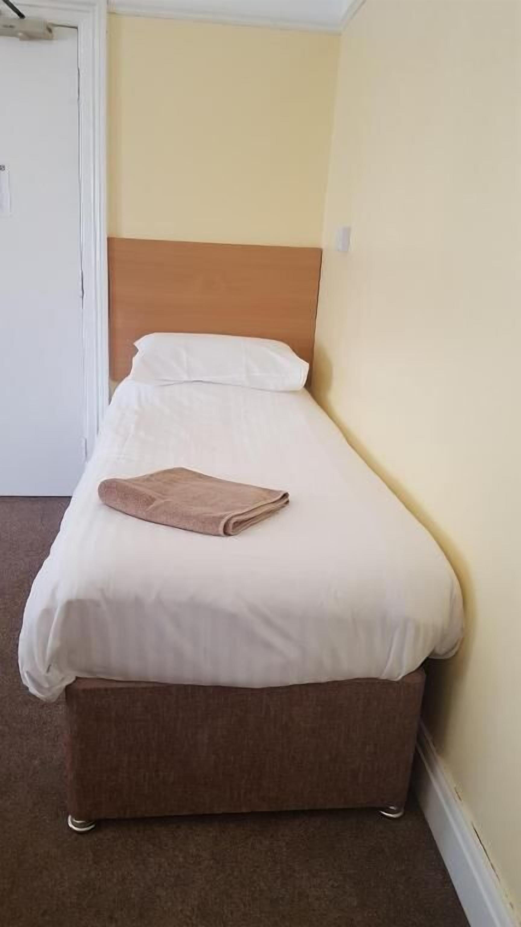 A small, single-bed room with a brown headboard, white bedding, and a folded brown towel on the bed.
