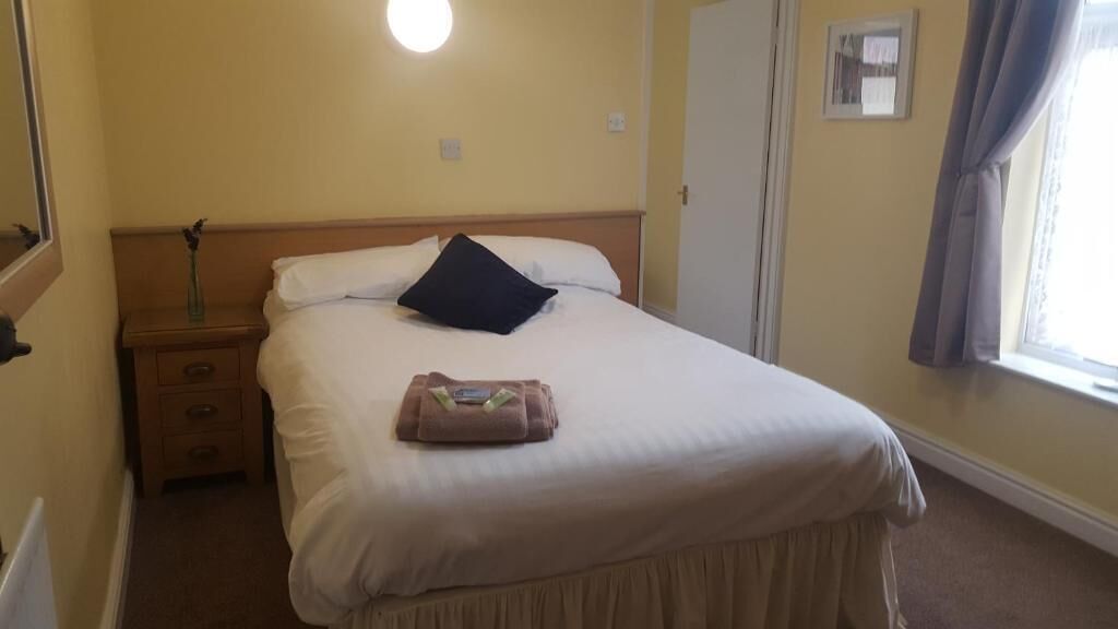 Una habitación de hotel con cama, mesita de noche, espejo, ventana con cortinas y un reloj de pared.
