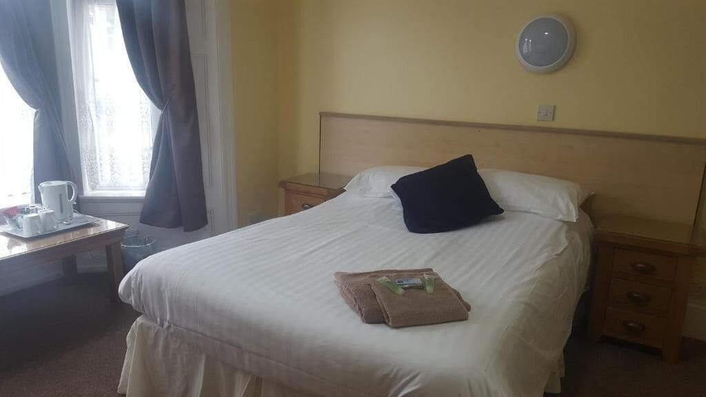 Une chambre d’hôtel comprenant un lit, une table de chevet avec une lampe, une fenêtre avec des rideaux et un luminaire fixé au mur.
