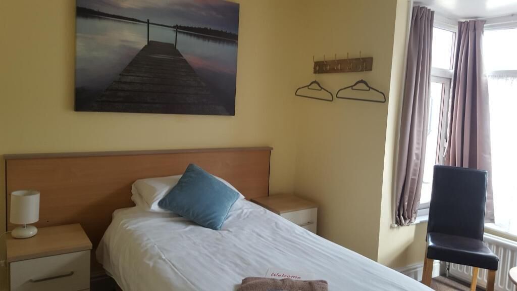 Ein Hotelzimmer mit Bett, Nachttisch, Lampe, Stuhl, einem Gemälde eines Piers und einem fenster mit Vorhängen.