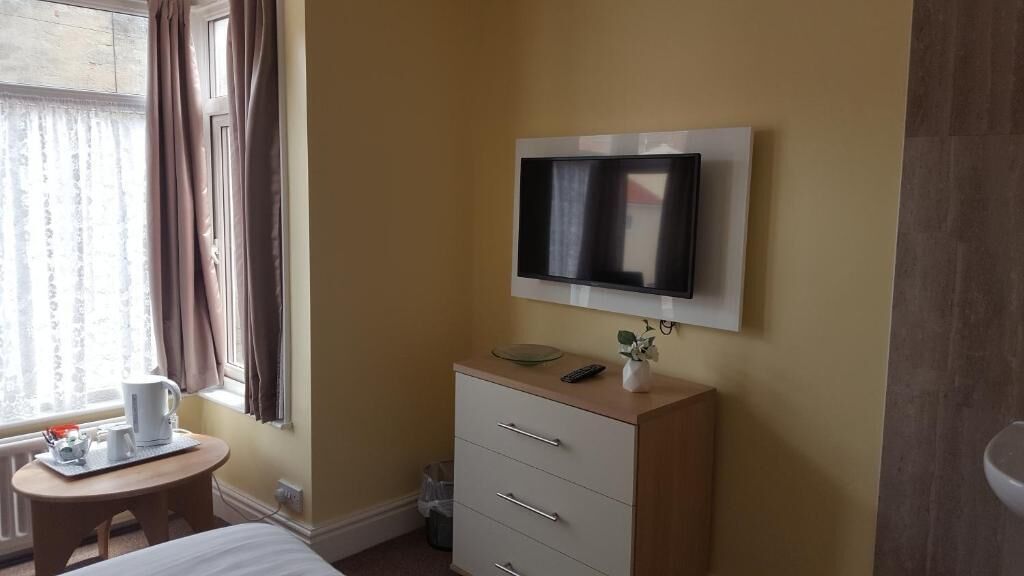 Hotelkamer met een bed, nachtkastje met koffiezetapparaat, een aan de muur bevestigde televisie, een klein kastje en een raam met gordijnen.