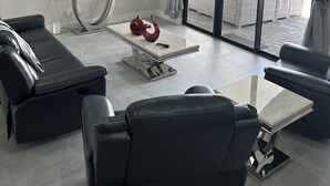 Een moderne woonkamer met een plafondventilator, een flatscreentelevisie, zwarte leren banken, een glazen salontafel en uitzicht op een terras.