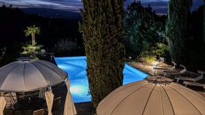 Une piscine entourée de chaises longues et de parasols, bordée de grands arbres et surplombant un ciel nocturne.