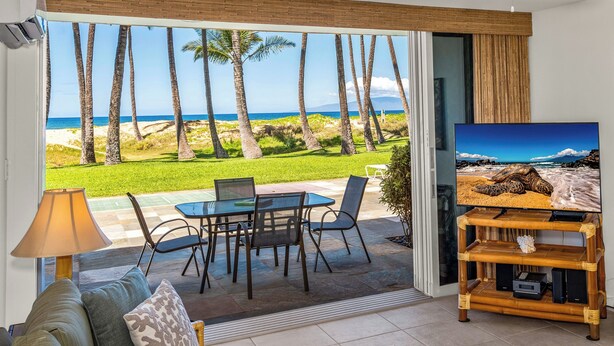 Ein Wohnzimmer mit Blick auf den Strand, einem Fernseher und einem Essbereich mit Stühlen und einem Tisch.