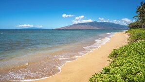 Ein Strand mit klarem, blauem Wasser, sandigem Ufer und einem Berg im Hintergrund.