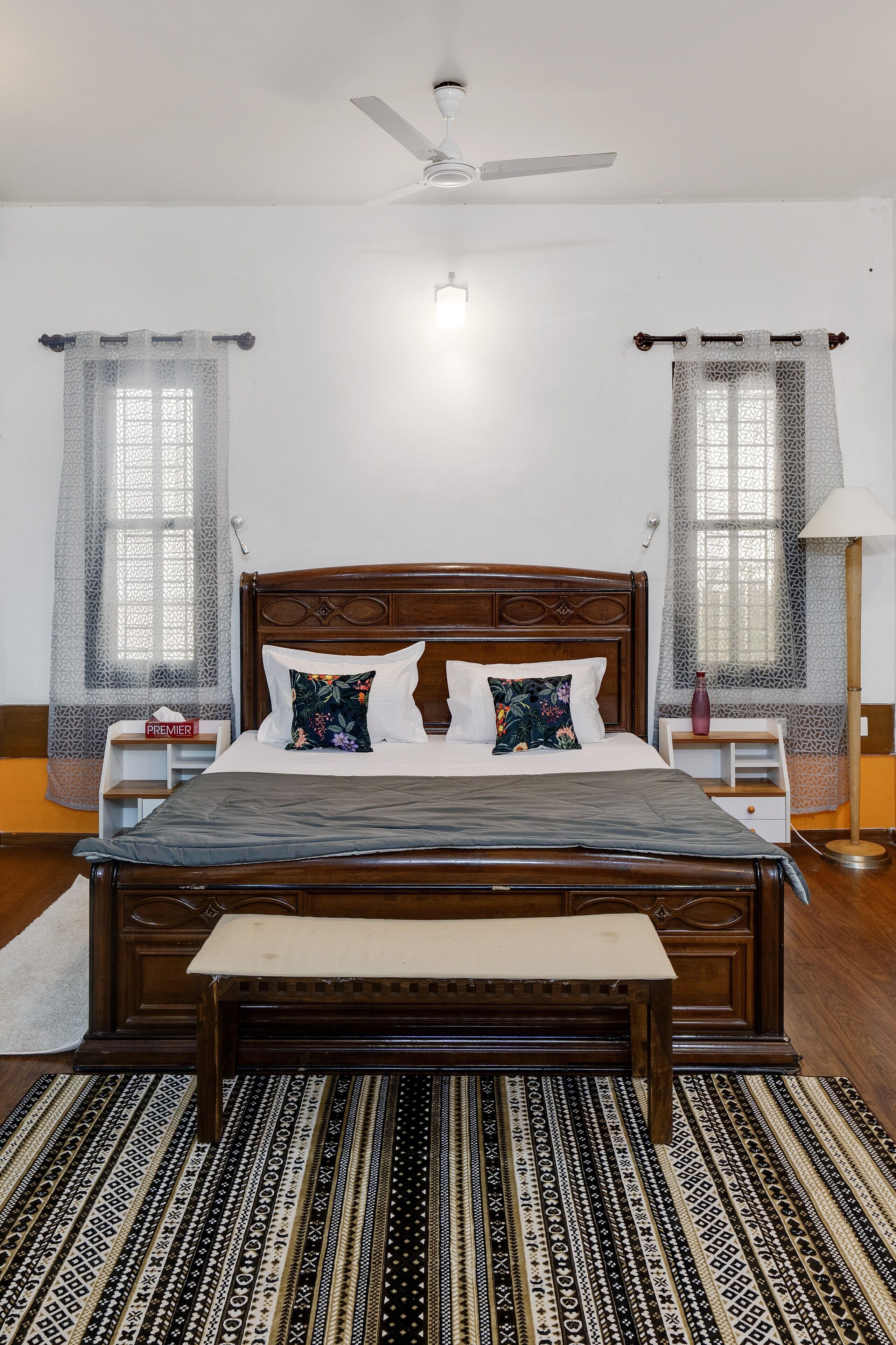A neatly made bed with a wooden headboard, a bedside table with a lamp, and a large window with a curtain.