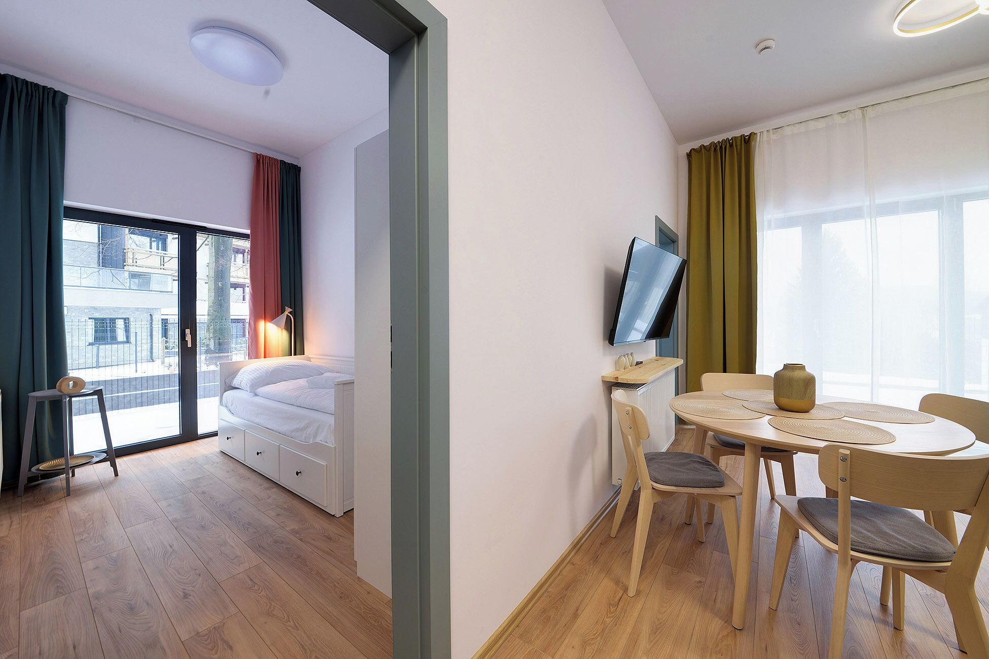 A modern room with a bed, a TV, a dining table with chairs, and a view of the outside.