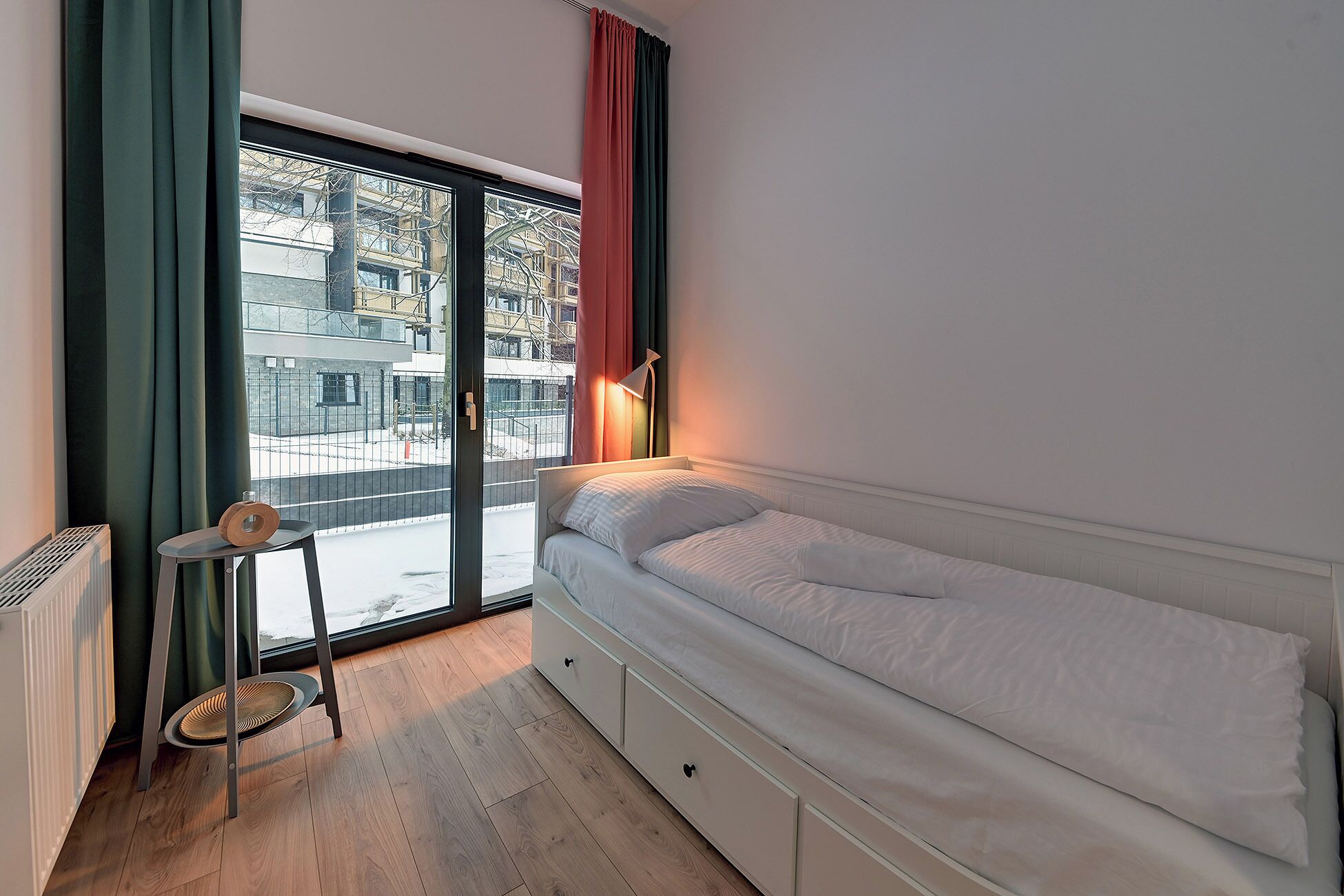 A small bedroom with a single bed, a chair, a radiator, and a window with curtains.