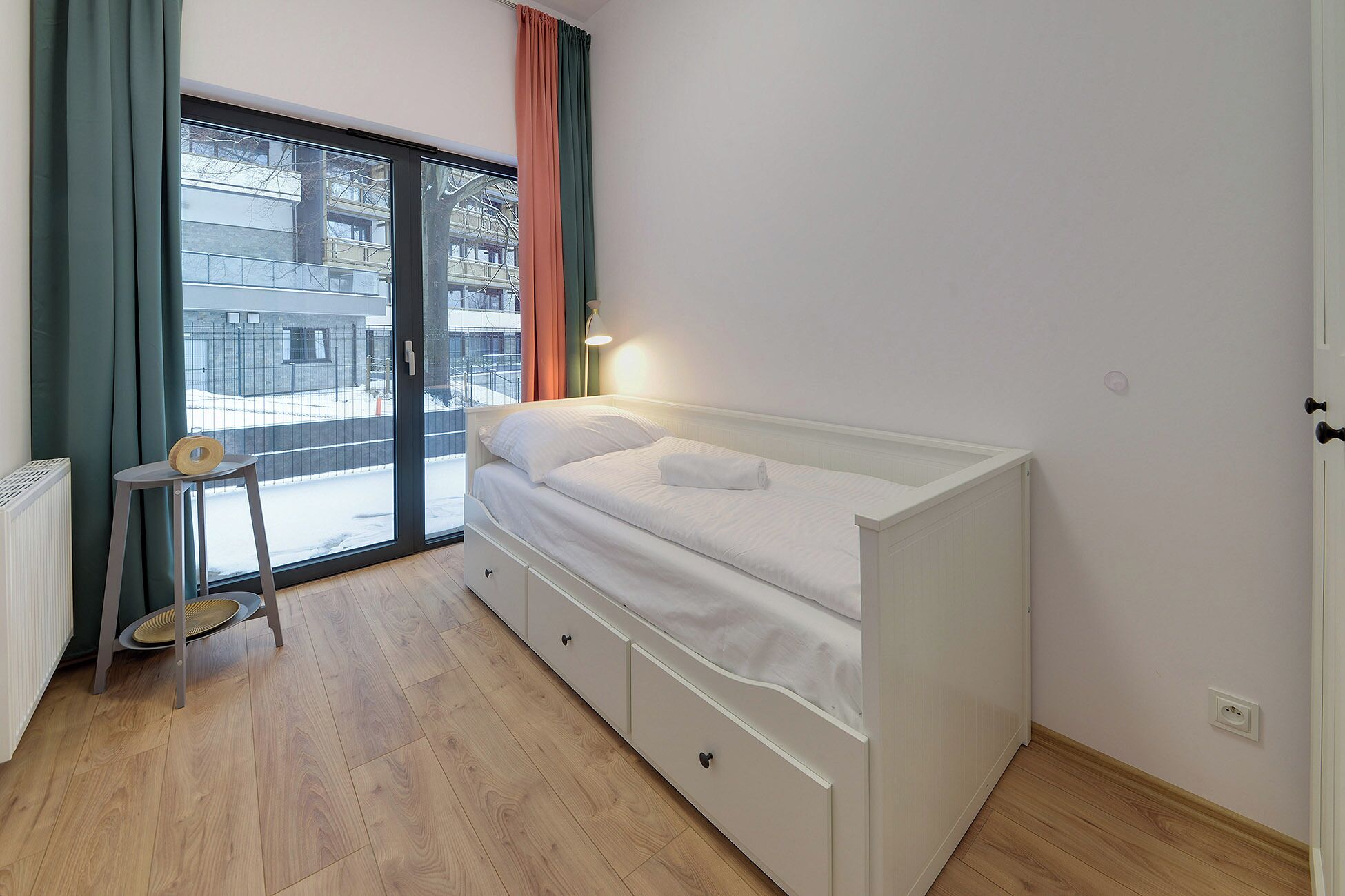 Een slaapkamer met een enkel bed, een kleine tafel met stoel, een radiator en een raam met gordijnen.