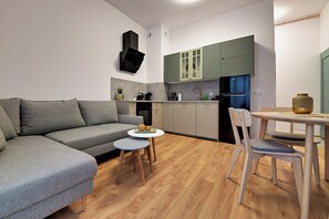 Un ambiente compatto con un divano grigio, un piccolo tavolino rotondo, un tavolo da pranzo con sedie e una zona cottura con pensili verdi.