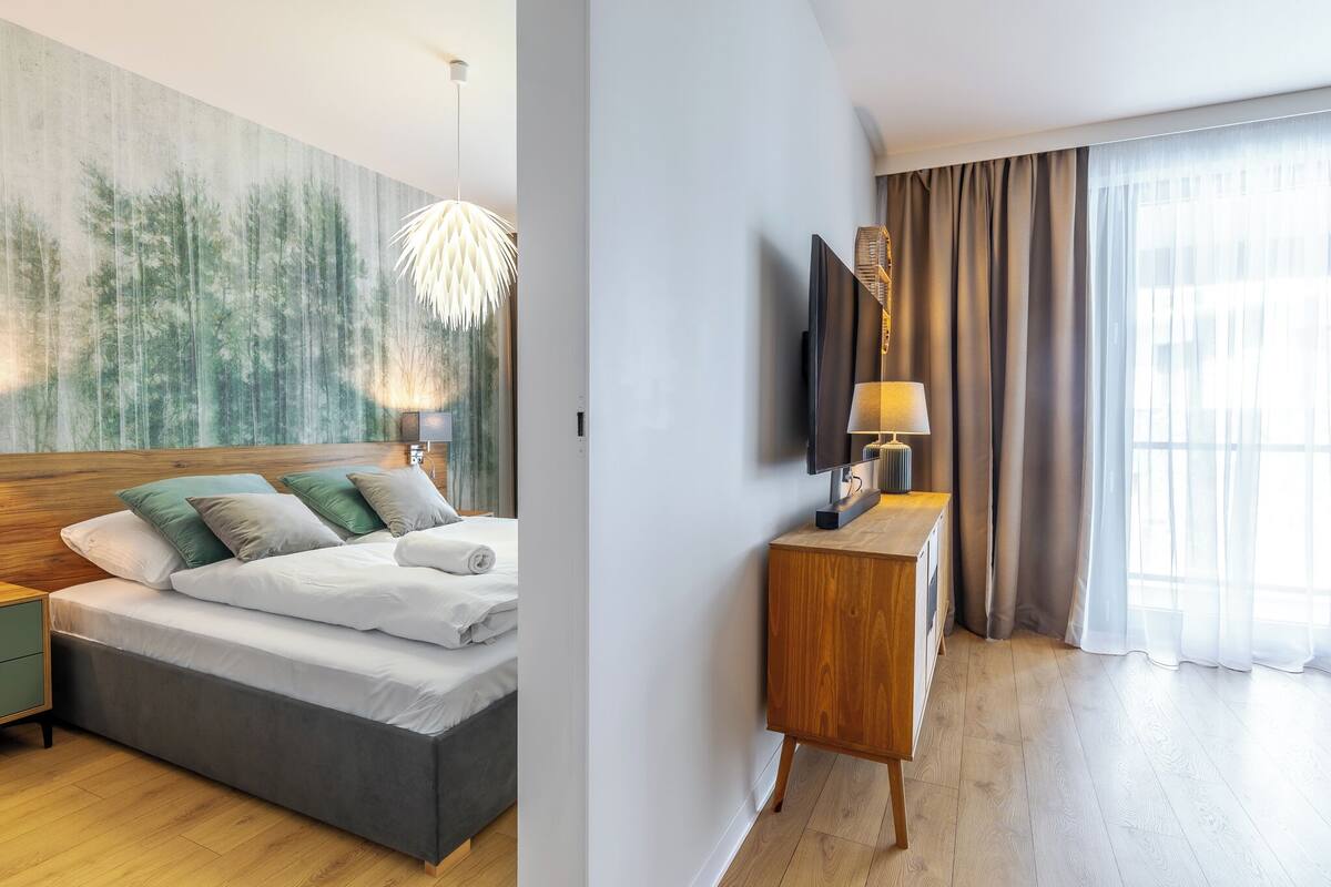 A modern hotel room with a large bed, a wooden headboard, a nightstand, a wall-mounted TV, a wooden console table, and a large window with curtains.
