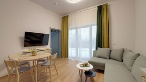 A modern living room with a sofa, a dining table with chairs, a flat-screen TV mounted on the wall, and large windows with sheer curtains. - RentPlanet - Źródło Izery (Swieradow-Zdroj)