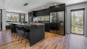 A modern kitchen with a large island, black cabinetry, and a stainless steel refrigerator. - Ski Saint-Come | Spa and Sauna | Le TAMARÄK (Lanaudière)