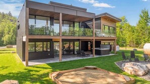 A modern two-story house with large glass windows, a covered outdoor seating area, and a well-maintained lawn. - Ski Saint-Come | Spa and Sauna | Le TAMARÄK (Lanaudière)
