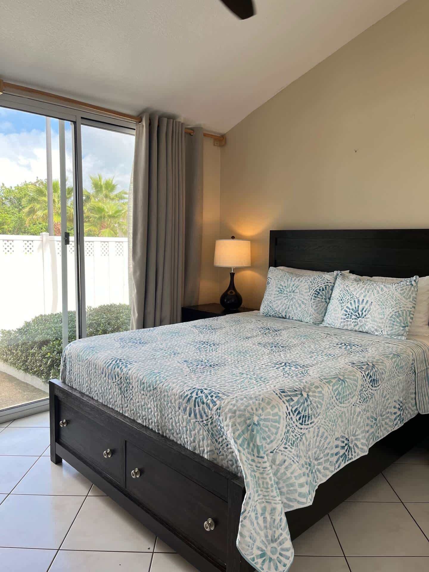 A bedroom with a bed, a nightstand, a lamp, and a view of palm trees through a sliding door.