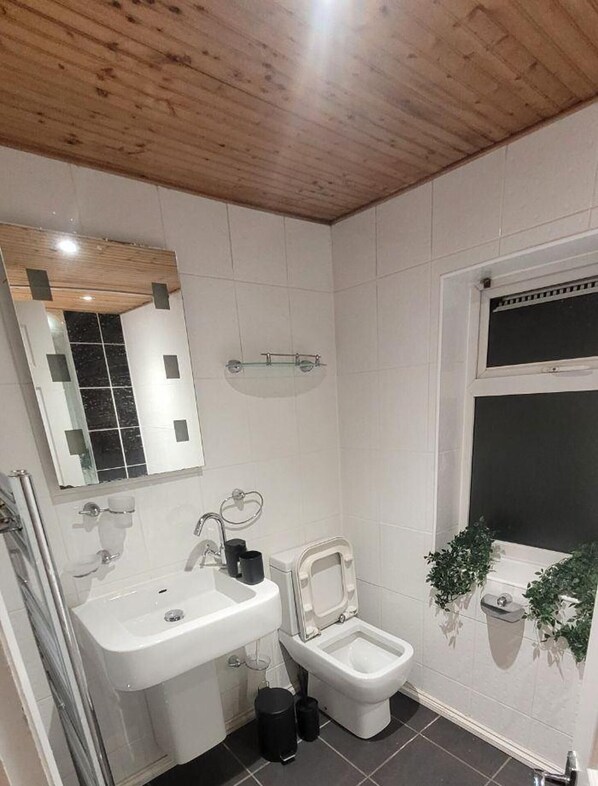 A bathroom with a wooden ceiling, white tiles, a sink, a toilet, and a window with a plant.