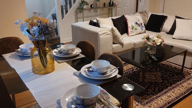 Un comedor con una mesa dispuesta para dos personas, un florero con flores y una escalera al fondo.