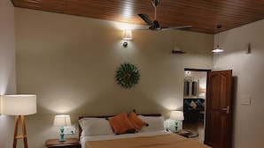 Un dormitorio con cama, dos mesitas de noche con lámparas, ventilador de techo, reloj de pared y puerta que da a otra habitación.