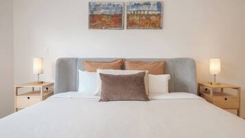 A neatly made bed with a grey headboard, two bedside tables with lamps, and two wall-mounted paintings.