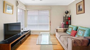 Ein modernes Wohnzimmer mit Flachbildfernseher, Glastisch und einem beigefarbenen Sofa mit Zierkissen.