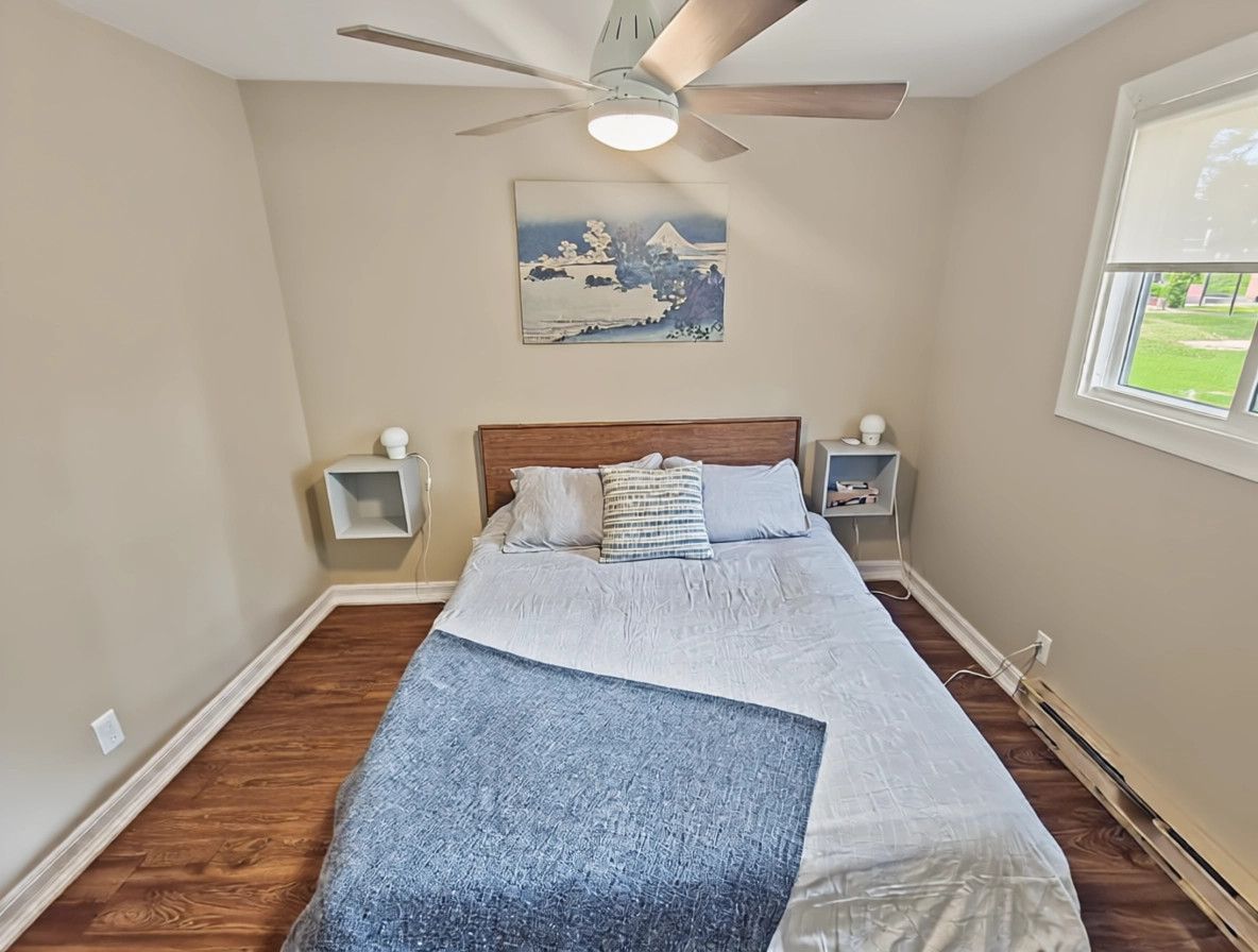 A bedroom with a bed, a ceiling fan, a nightstand, a lamp, and a window with a view of the outdoors.
